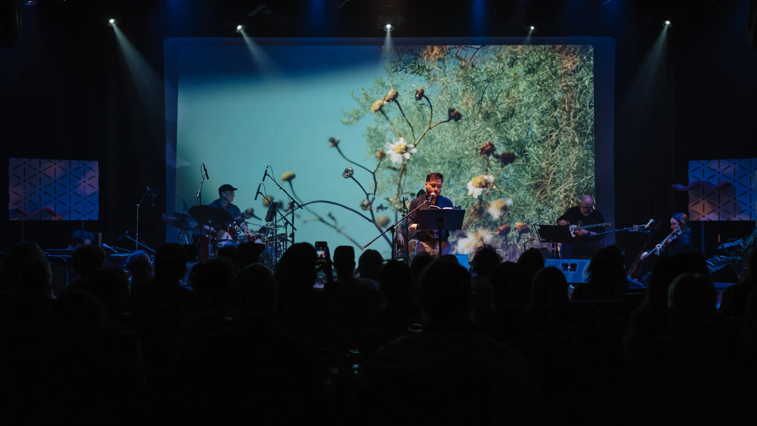A man in front of a backdrop image of flowers, speaking into a microphone to an audience. 
