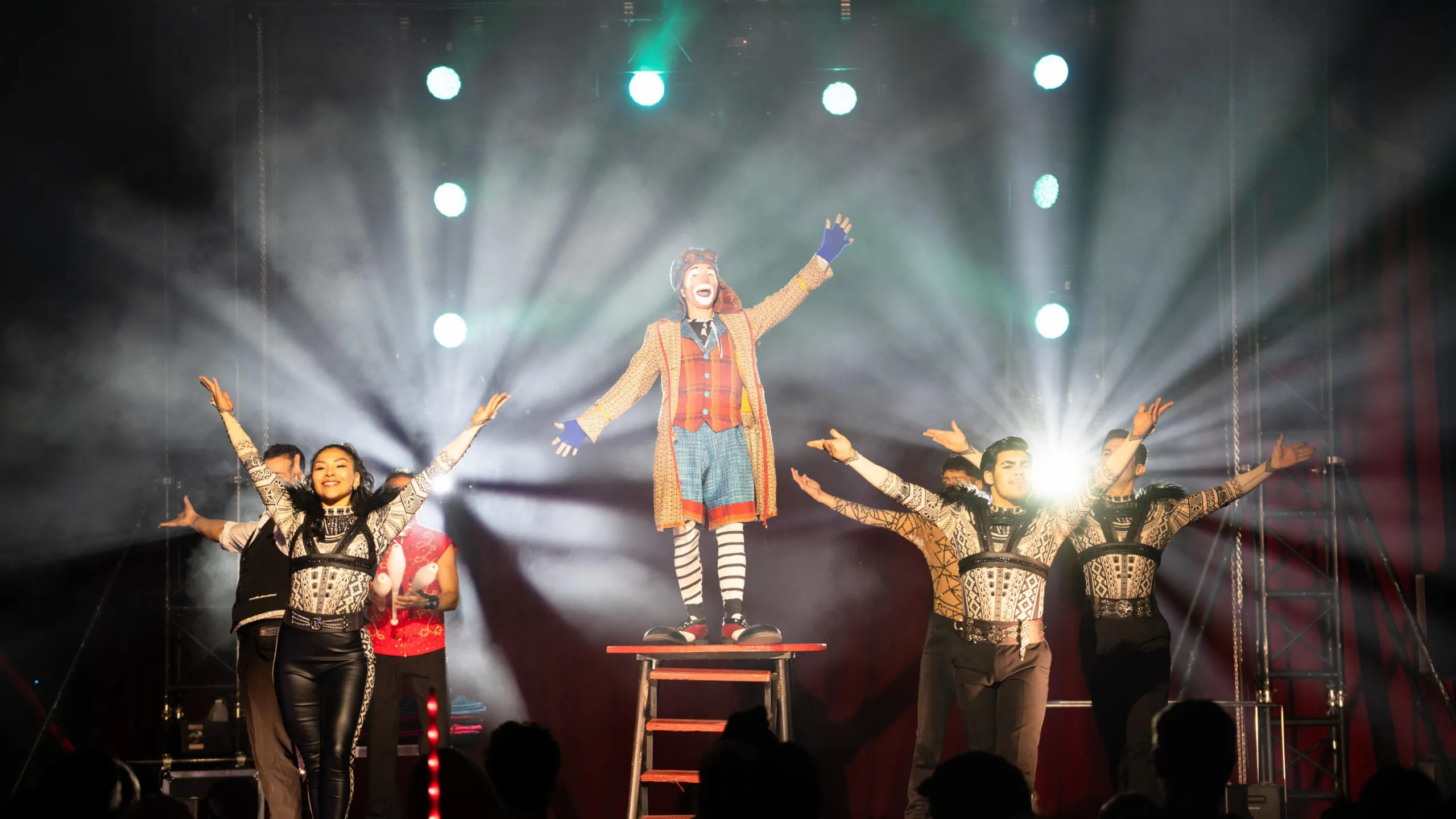 A clown and his assistants stand on a backlight stage, waving to an audience. 