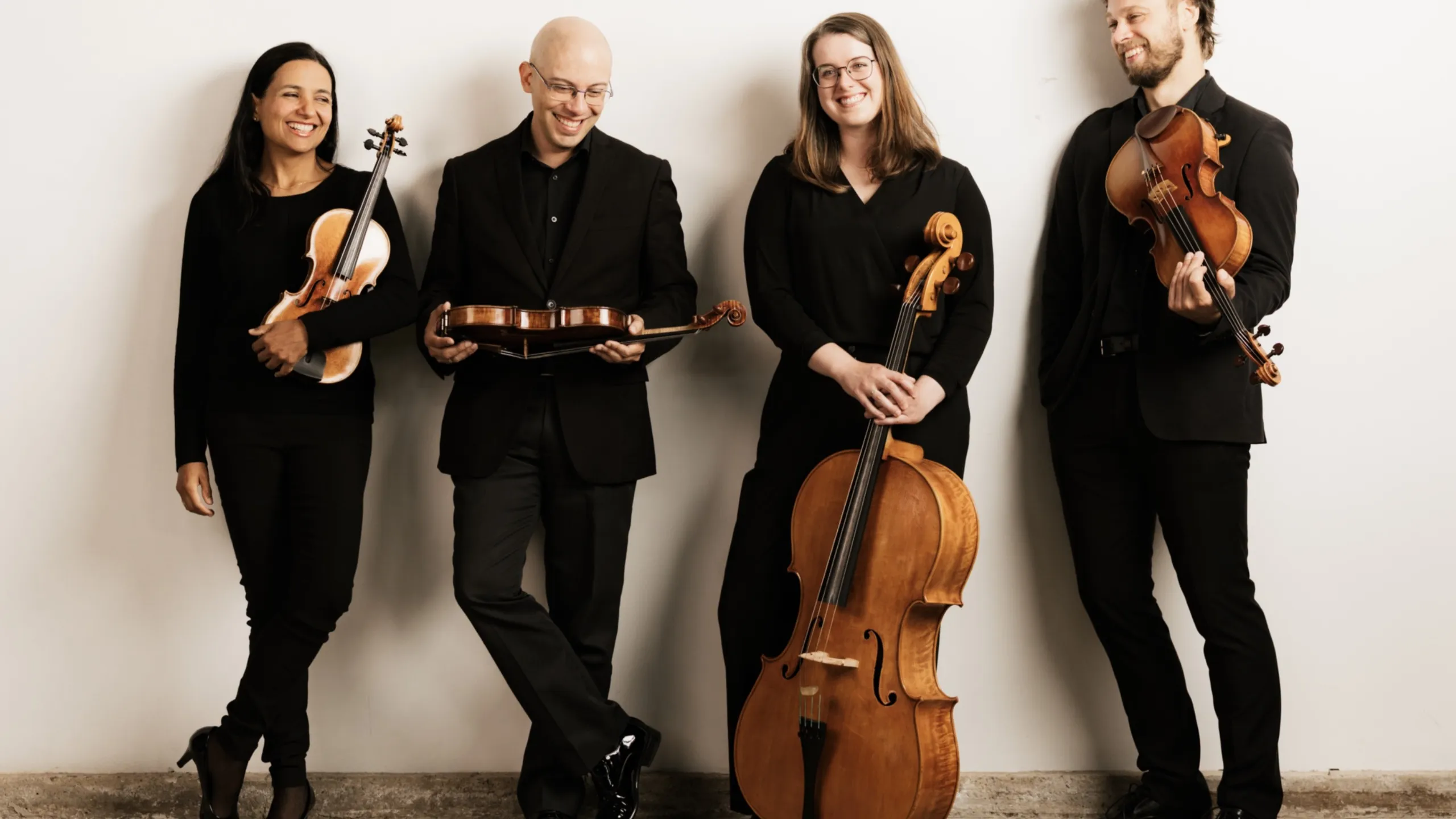 Four musicians holding their stringed instruments (three violins and a cello) and smiling. 