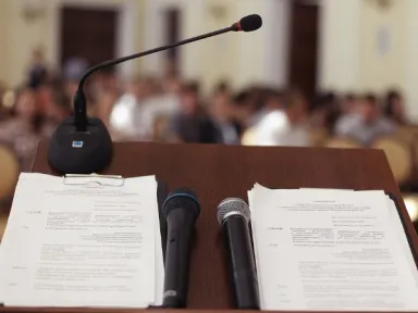 Podium with papers and two handheld microphones; gooseneck mic facing a blurred audience.
