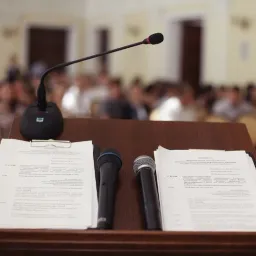 Podium with papers and two handheld microphones; gooseneck mic facing a blurred audience.